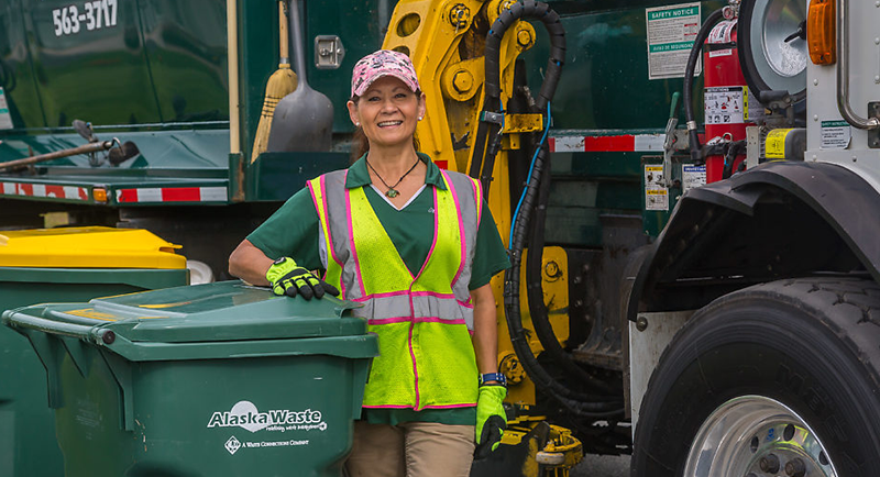 Closeup photo of Alaska Waste employee standing between a truck and a bin.